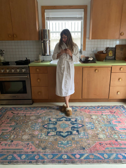Person standing in a kitchen with wooden cabinets and a patterned rug.
